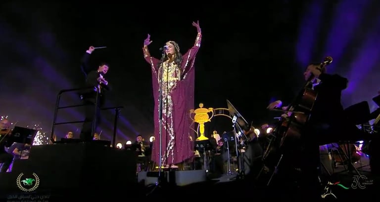 Performer on stage wearing a sparkly purple outfit with arms raised, surrounded by band members and stage lighting