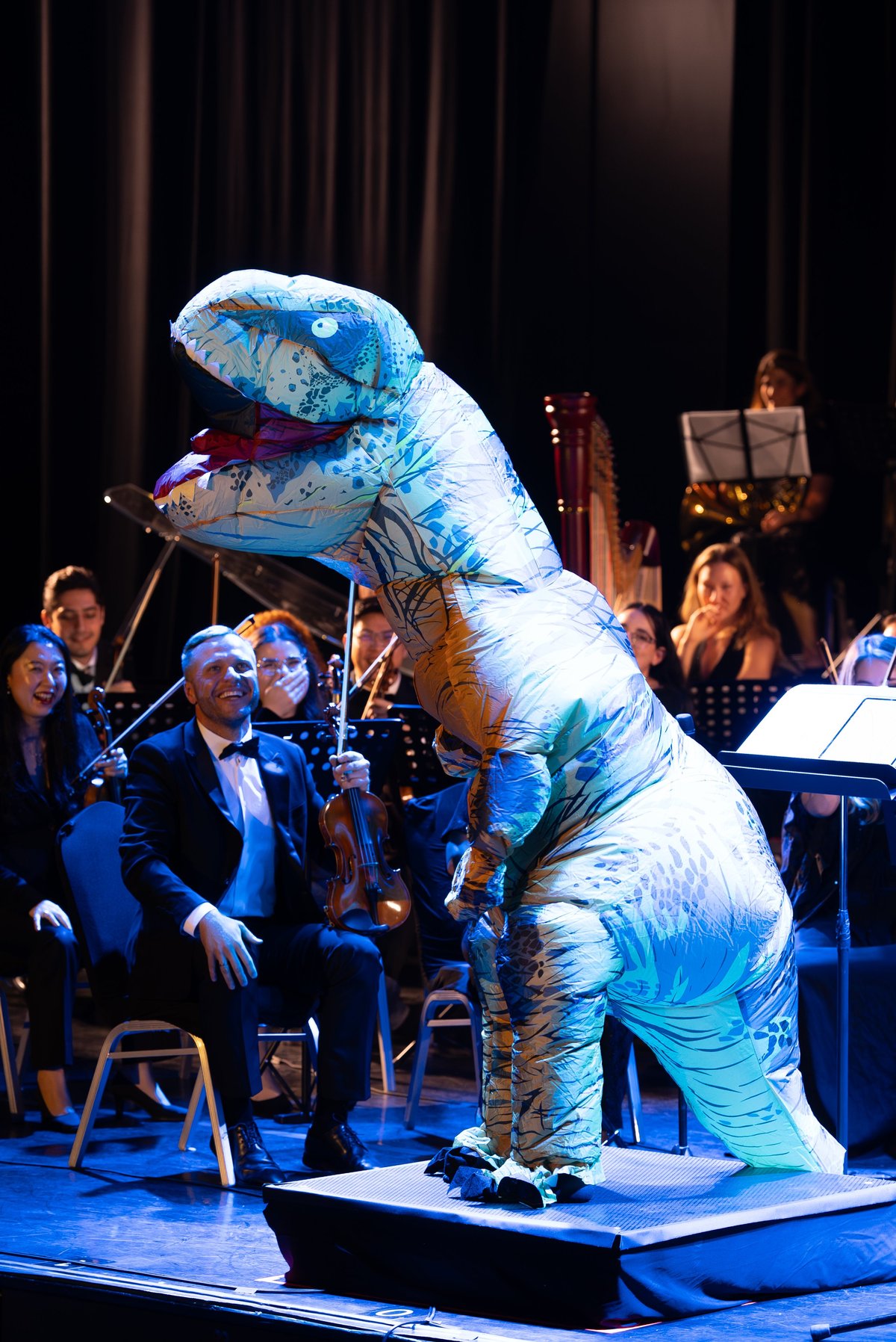 Person in blue and white dinosaur costume on stage interacting with orchestra musicians during a concert performance
