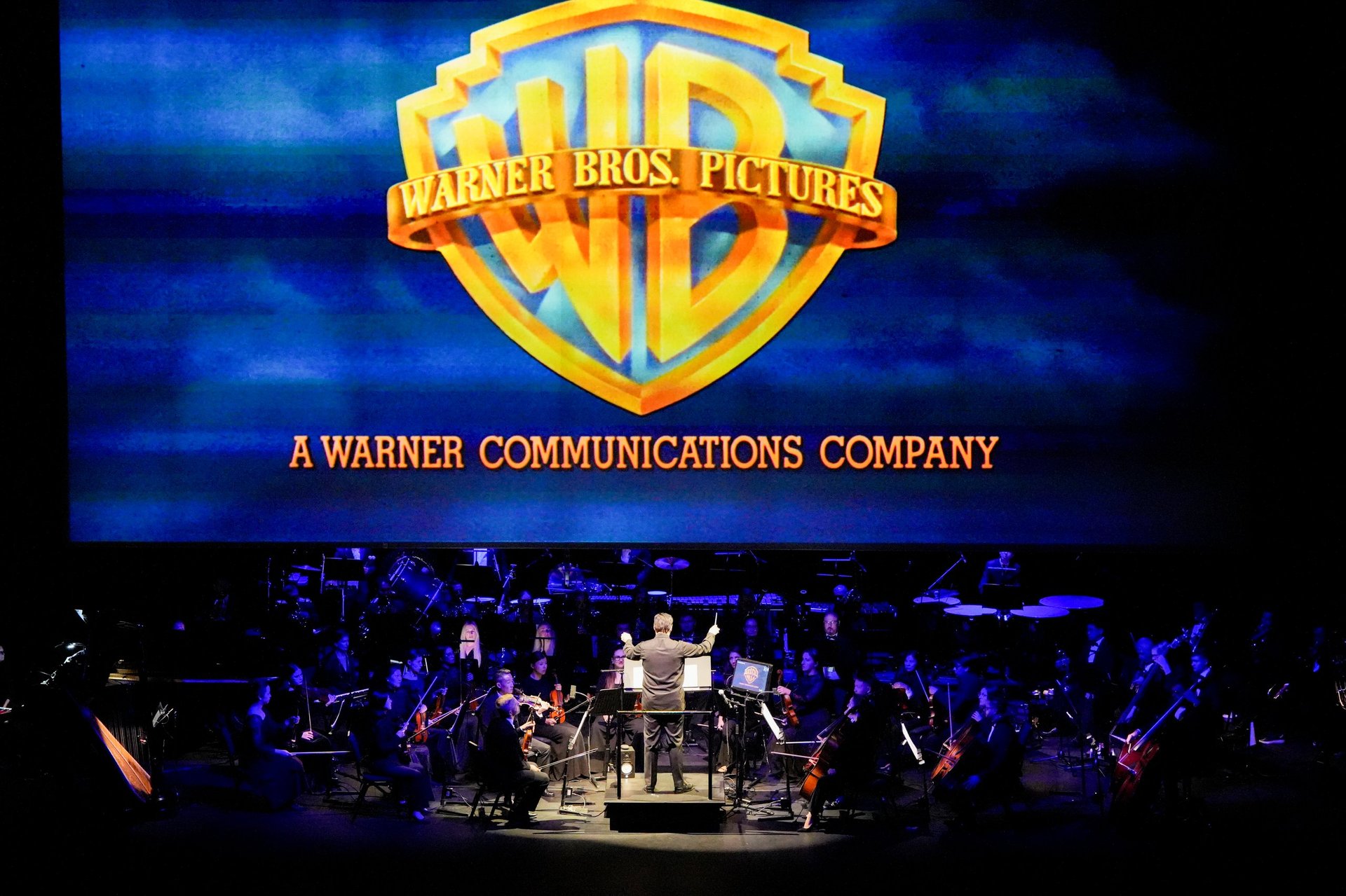 Warner Bros. logo on screen with orchestra performing on stage below during live concert